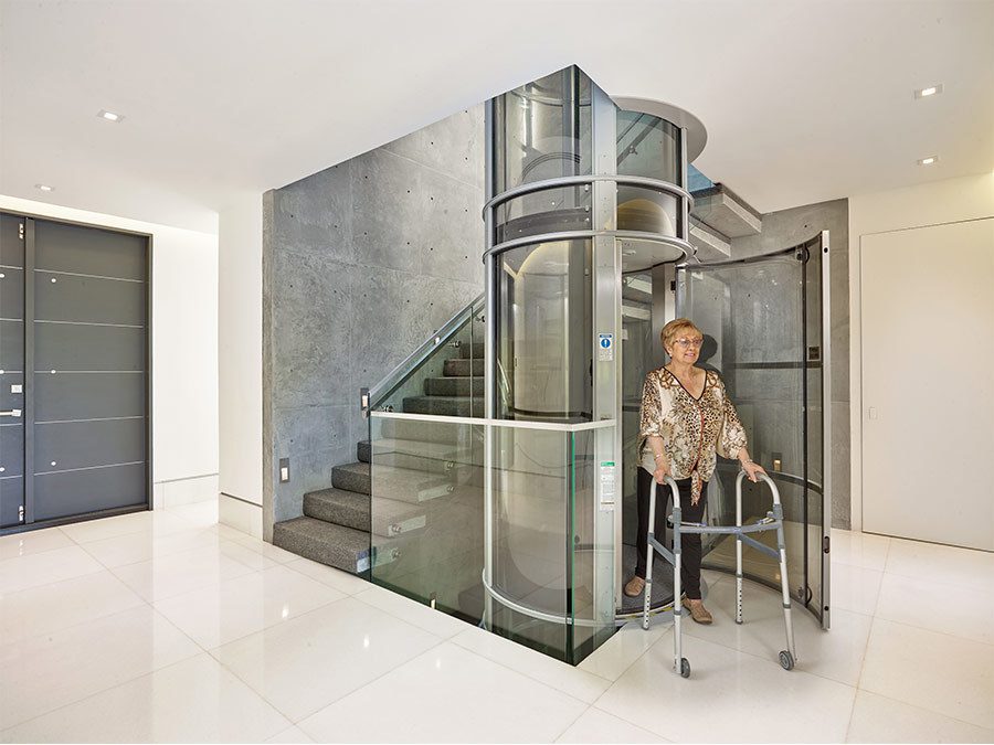 Elderly woman beside glass elevator in home.